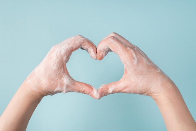 Concetto di lotta al coronavirus. foto ritagliata ravvicinata delle mani femminili che mostrano la forma del cuore con due mani in schiuma di gel di sapone su sfondo di colore blu pastello