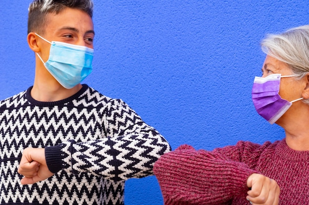 Coronavirus. Greetings with elbows for a white-haired grandmother and her teenage grandson wearing a surgical mask due to coronavirus, smiling and looking each other. New normal concept