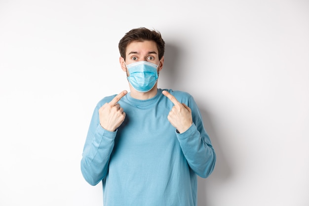 Coronavirus, health and quarantine concept. Confused guy pointing at his medical mask and looking surprised at camera, standing over white background.