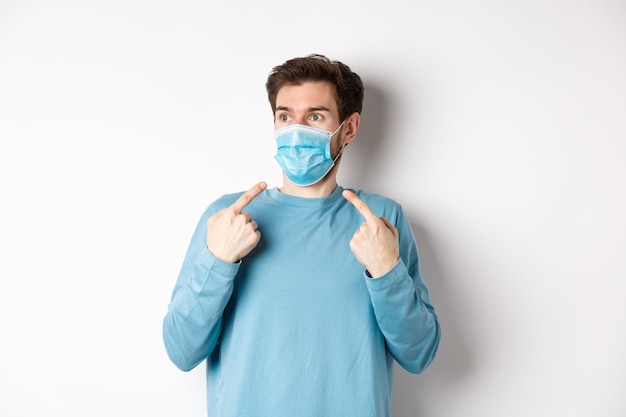 코로나 바이러스, 건강 및 검역 개념. 혼란 스 러 워 젊은 남자 얼굴에 의료 마스크를 가리키고 흰색 배경 위에 서있는 왼쪽을보고.