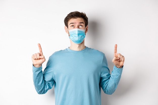 コロナウイルス、健康と検疫の概念。好奇心旺盛な男は、白い背景の上に医療マスクに立って、上にバナーを読んで、指を上に向けて見ています。