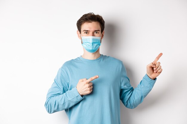 Coronavirus, health and quarantine concept. Handsome caucasian man in face mask pointing fingers right, showing advertisement, standing over white background.