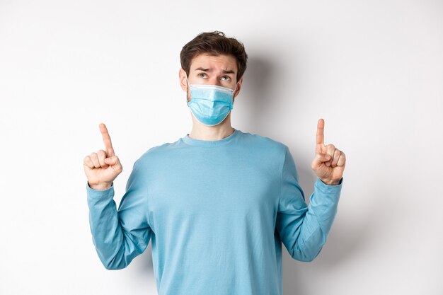 Coronavirus, health and quarantine concept. Skeptical and doubtful man in medical mask pointing, looking up, feeling indecisive, standing over white background.