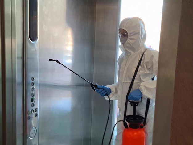 Coronavirus infection and flu paramedic in protective mask and suit disinfects elevator with spray bottle