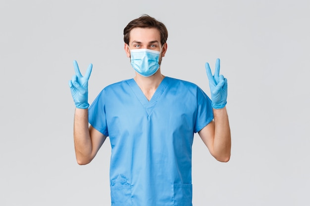 코로나바이러스 발병, 질병과 싸우는 의료 종사자, 병원 개념. 파란색 스크럽과 장갑, 의료 마스크를 쓴 친절하고 낙관적인 의사, 대유행 covid-19 동안 긍정적으로 웃고 있습니다.