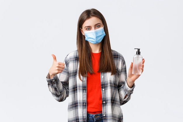 Coronavirus outbreak, leisure on quarantine, social distancing and emotions concept. Cheerful pleased young girl recommend hand-sanitizer brand, thumb-up, wear medical mask