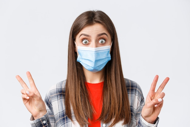 Coronavirus outbreak, leisure on quarantine, social distancing and emotions concept. Close-up of excited and thrilled cute woman in medical mask, showing peace sign or quotes, stare impressed.