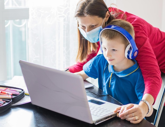 Coronavirus Outbreak. Lockdown and school closures. Mother helping her son with face mask studying online classes at home. COVID-19 pandemic forces children and teachers online learning.