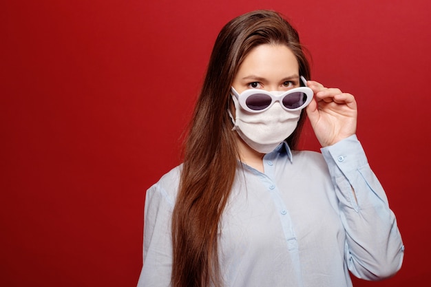 Coronavirus pandemic, close-up portrait of young woman on red background in protective medical mask and sunglasses