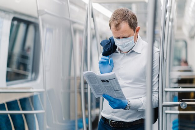 Coronavirus pandemic, Covid-19, quarantine measure. Serious adult man in white shirt, holds black jacket, concentrated in newspaper, wears protective mask and gloves, poses at metro carriage