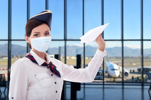 Coronavirus, pandemic, vacation and travel concept - young beautiful stewardess in medical mask posing with paper plane in airport