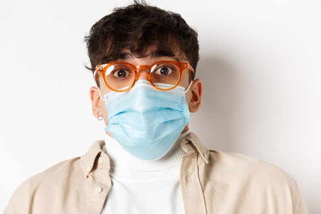 Coronavirus pandemie en gezonde mensen concept. Verrast man in bril en gezichtsmasker wenkbrauwen optrekken, vol ontzag staren, staande op een witte muur.