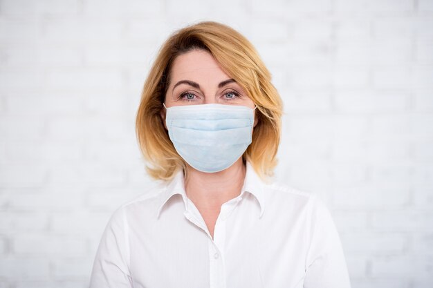 Coronavirus, pandemie, quarantaine en gezondheidszorgconcept - close-up portret van volwassen vrouw met medisch masker over witte bakstenen muur