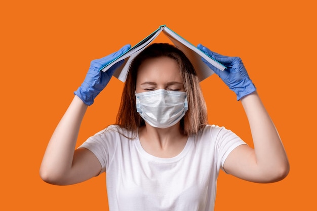 Coronavirus paniek Infectierisico Angstige vrouwelijke student met gezichtsmasker handschoenen die hoofdbedekking beschermen met open boek geïsoleerd op oranje Quarantaine hygiënemaatregelen COVID19 pandemie Veilige studie