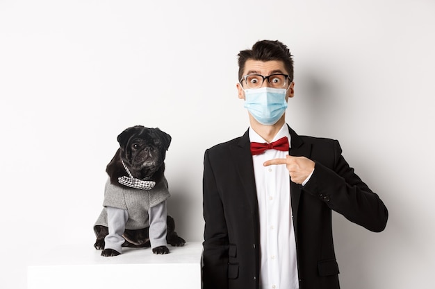 Coronavirus, pets and celebration concept. Amazed young man in face mask and suit pointing at cute black dog sitting near owner in party outfit, white background