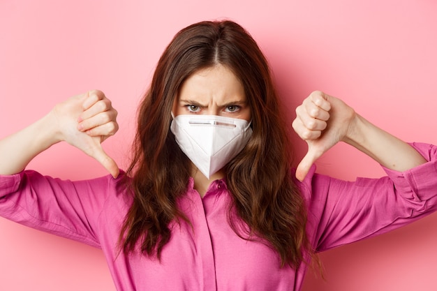 Coronavirus, preventive measures and health concept. Close up portrait of angry young woman in respirator frowning, showing thumbs down with furious face, pink wall.