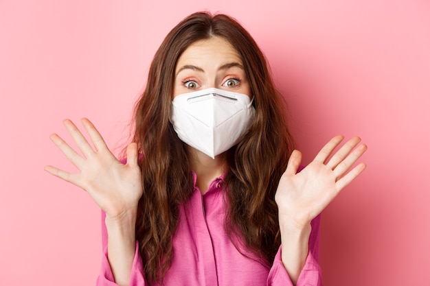 Coronavirus, preventive measures and health concept. Close up portrait of surprised and excited woman in respirator, raising hands up and gasping amazed, pink wall.