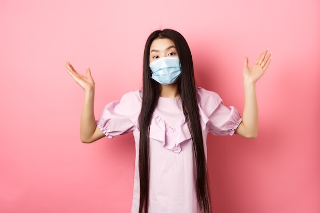 Photo coronavirus, quarantine and lifestyle concept. clueless asian girl in face mask shrugging shoulders unware, know nothing, standing on pink background.