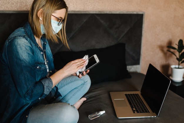 Coronavirus. Sanitizer. Woman working, wearing protective mask in quarantine. Cleaning hands mobile phone to eliminate germs antibacterial spray. Disinfecting wipes for cleaning phone. Stay at home.