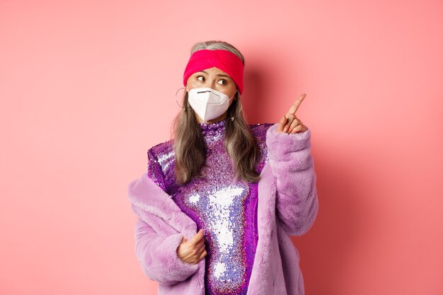 Coronavirus and shopping concept. Sassy elderly asian woman in respirator and glittering shiny dress, pointing at upper right corner and look doubtful, pink background.