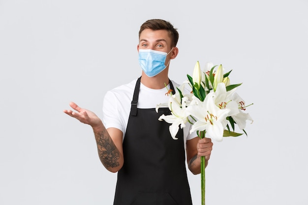 Coronavirus, social distancing business in covid-19 pandemisch concept. Vriendelijke charismatische verkoper in schort en medisch masker bereidde een prachtig boeket lelies, pratend met de klant in de bloemenwinkel