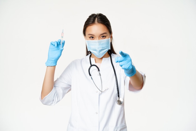 Coronavirus vaccination. Asian physician, doctor holding vaccine syringe and pointing at camera patient, vaccinating campaign, white background