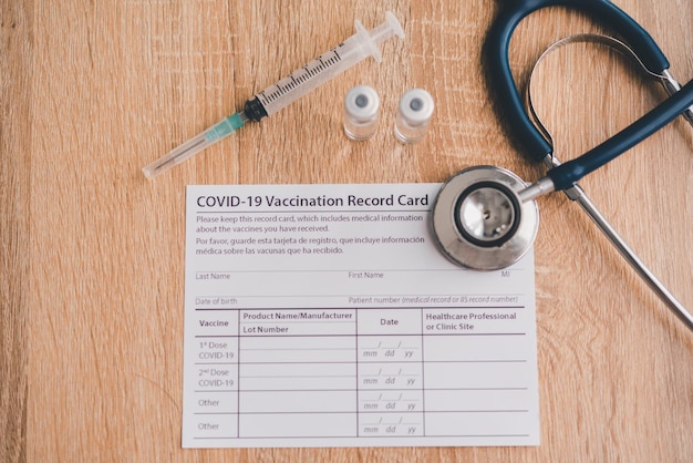 Coronavirus vaccination record card is placed on a wooden floor with a vaccine syringe and a stethoscope.