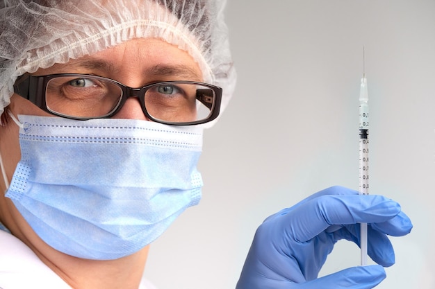 Концепция вакцины против коронавируса. Медик, врач или медсестра с флаконом с вакциной covid-19 и шприцем. Зрелая европейская женщина-техник в маске, очках, одноразовой шляпе, перчатках и белом защитном платье.