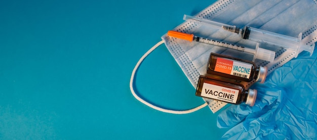 Фото Коронавирусная вакцина в бутылках на синем фоне с копией пространства флаконы с лекарством для шприца covid19, медицинская маска для лица и перчатки, вид сверху баннер концепции иммунизации