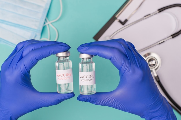 코로나바이러스 백신 테스트 검사 개념. 파란색 청록색 배경에 격리된 마스크 청진기와 클립보드가 있는 유리병을 들고 있는 의사의 머리 위 사진