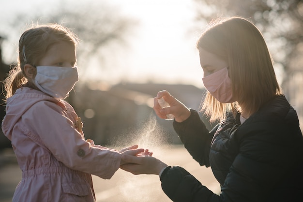 Coronavirus. Vrouw in een beschermend masker gebruik spray ontsmettingsmiddel op handen kind op straat. Preventieve maatregelen tegen Covid-19-infectie. Antibacteriële handwasspray. Ziekte bescherming.