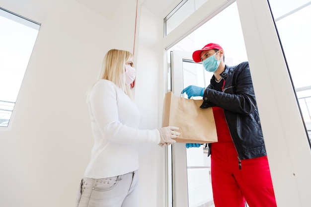 Coronavirus. Vrouw met een medisch masker en rubberen handschoenen die binnen een pakket ontvangt van een bezorger. Viruspreventie en protocollen. Blijf thuis. Bezorgservice. Desinfecteren van het pakket.