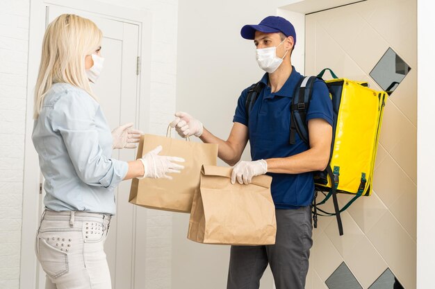 Coronavirus. Woman wearing a medical mask and rubber gloves receiving a package from a delivery man indoors. Virus prevention and protocols. Stay at home. Delivery service. Disinfecting the package.