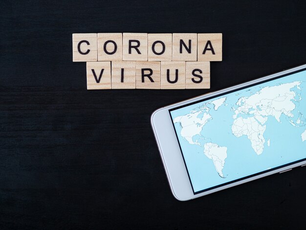 Coronavirus word gemaakt van hout blok en wereldkaart op smartphone scherm. Coronavirus tekst op dramatische sfeer zwarte houten tafel. Coronavirus concept bovenaanzicht, kopie ruimte