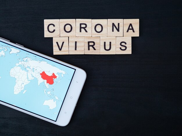 Coronavirus word made of wood block and world map on smartphone screen. Coronavirus text on dramatic atmosphere black wooden table. Coronavirus concept top view, copy space