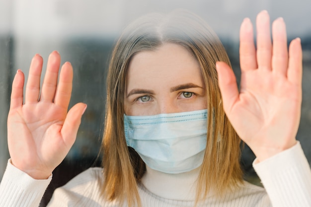 Coronavirus. Zieke vrouw die het masker draagt dat van het beschermingsgezicht door het venster kijkt