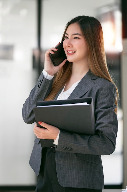 Corporate businesswoman holding file folder and talking on mobile phone standing in office