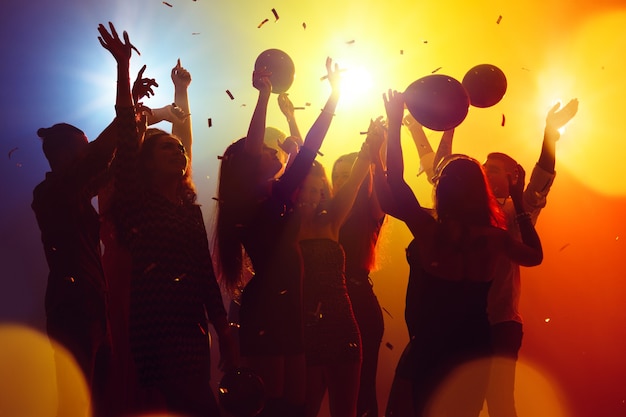 Corporate. A crowd of people in silhouette raises their hands on dancefloor on neon light background. Night life, club, music, dance, motion, youth. Yellow-blue colors and moving girls and boys.