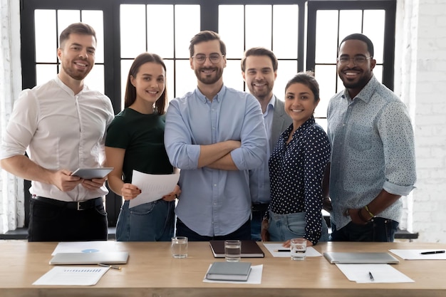 Фото Корпоративное фото, улыбающиеся разнообразные сотрудники с уверенным руководителем в очках, стоящие в современном офисе и смотрящие в камеру, успешный основатель стартапа с членами команды