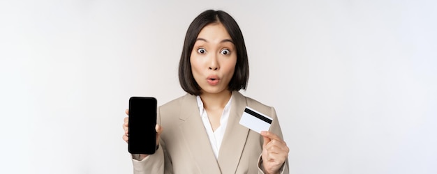 Corporate woman with happy enthusiastic face showing credit card and smartphone app screen standing in suit over white background