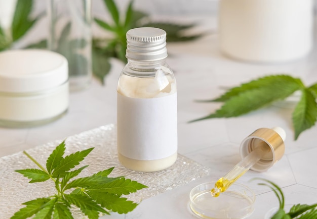 Cosmetic bottle with blank label near green cannabis leaves on white table Mockup