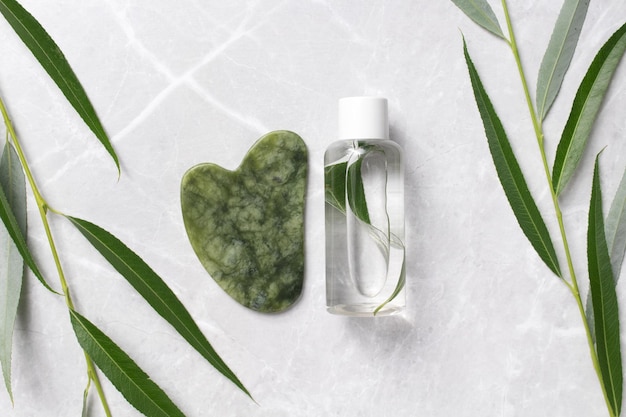 Cosmetic bottle with tonic water guasha with leaves on marble background Jade face massager for facial massage therapy Anti age lifting and toning treatment