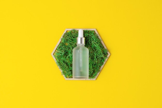 Cosmetic bottle with a wooden hexagon and moss on yellow background flat lay copy space