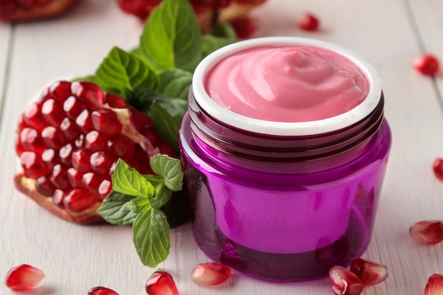 Cosmetic cream in a lilac jar and with fresh pomegranate on a white wooden table.