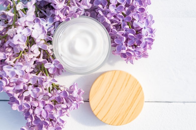 Cosmetic cream with fresh branches of purple lilac blossoms on light wooden background Dermatologic components for cosmetics Top view