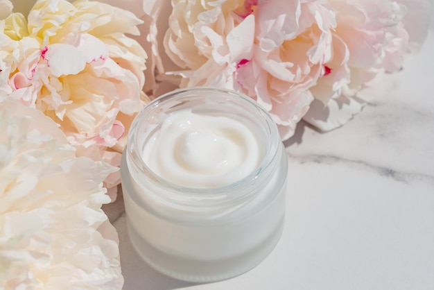 Cosmetic cream with fresh flowers of pink peony blossoms on light marble background Dermatologic components for cosmetics