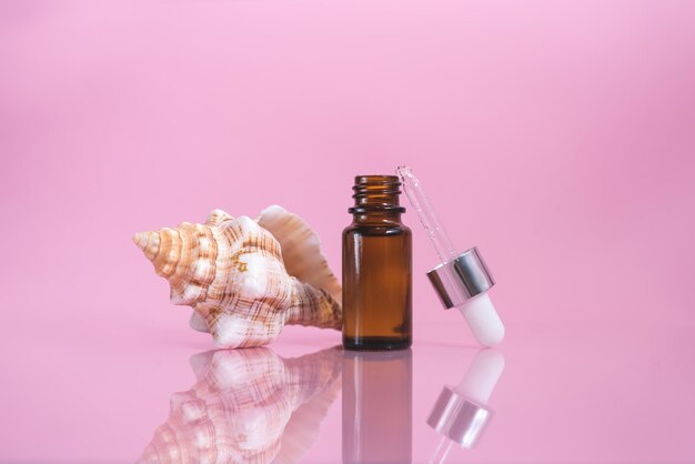 A cosmetic dropper bottle and sea shell on the pink background
