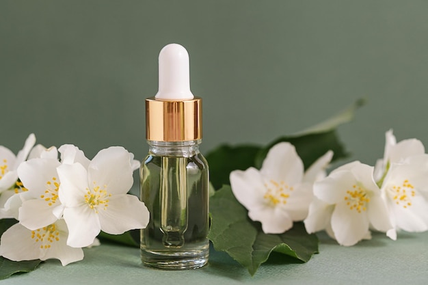 Cosmetic glass bottle for essential oils with jasmine flowers\
on green background spa natural serums dropper bottle mockup herbal\
medicine or cosmetics