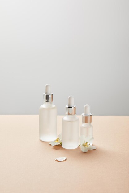 Cosmetic glass bottles and few jasmine flowers on beige isolated on grey