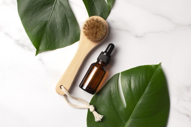 Cosmetic serum in brown glass bottle and body brush on a marble background with monstera palm leaf